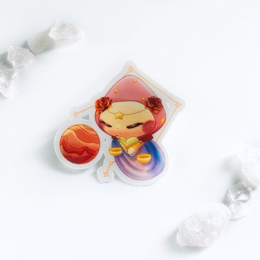 Cartoon character of a bunny in a strawberry hat wearing a beige outfit with gold bands. She is holding a gold balance and a flowing blue and purple clock under her. She is also wearing a golden star on her forehead and two red-golden roses. There is Venus next to her and the Libra constellation behind her. The background is white and there are small clear quartz.